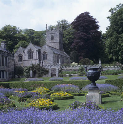View of the east end of the church and spring bluebells among the borders of the garden, with a bronze urn in the foreground. St Hydroc church is a parish church and not owned by the National Trust.